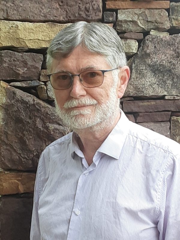 A man with gray hair and a beard is wearing a pin-stripe light-colored dress shirt and glasses, smiling, in front of a stone wall.