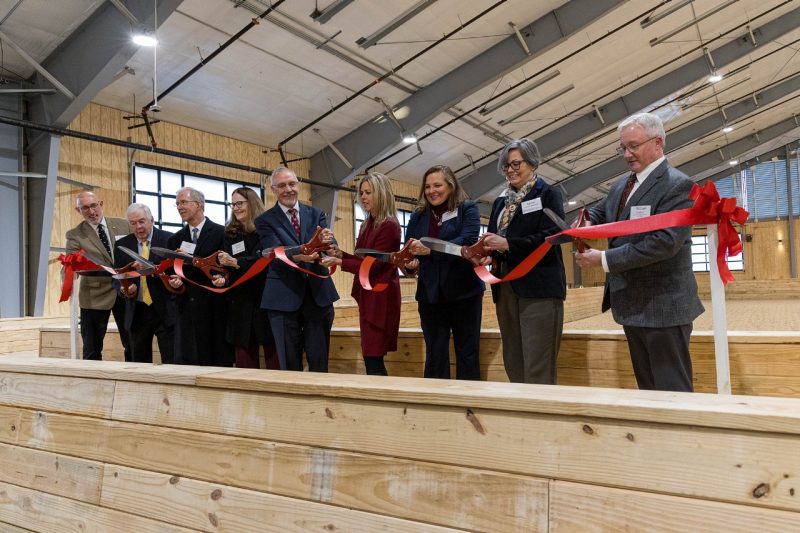 Group of people with large scissors, cutting a red ribbon in an indoor equine arena.