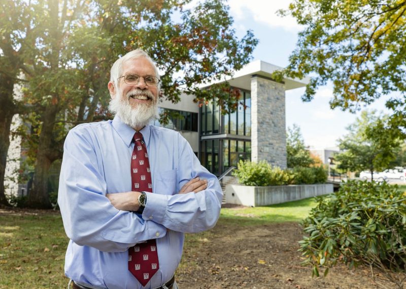 Man standing with his arms crossed with a collegiate building in the background.
